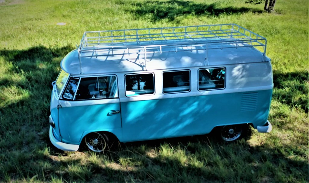 Overhead view of a restored vintage turquoise and white 1964 Volkswagen camper van parked on grass in a sunny field, with a large metal roof rack on top.