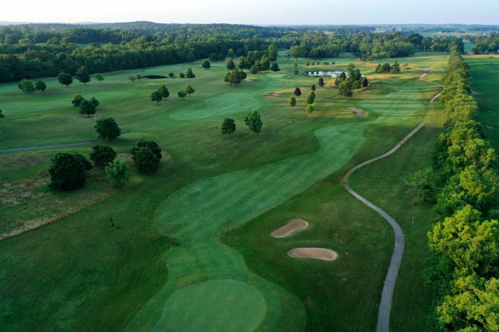 Aerial view of a lush green golf course with rolling fairways, scattered trees, sand bunkers, and small ponds, surrounded by dense forest and distant hills under a clear sky.